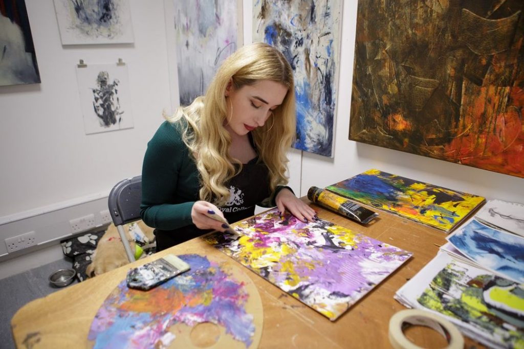 Kimberly Burrows, a woman with long blonde wavy hair, is sitting at a desk in her studio working on a painting